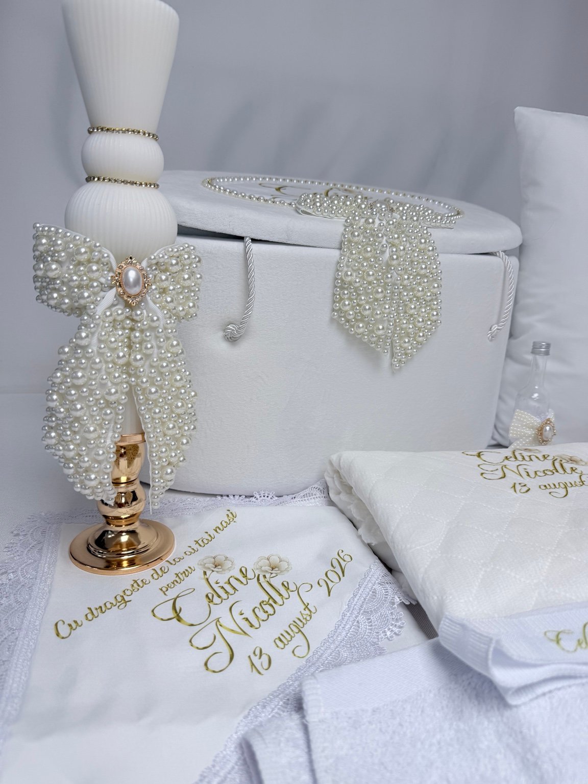 Detaliu al lumânării albe cu fundă din perle decorative și ornament oval auriu, lângă cufărul asortat