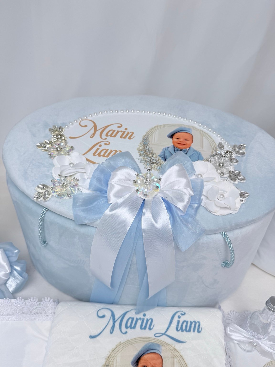 Cufăr bleu și lumânare canelată cu flori albe, funde și ornamente argintii, alături de textile personalizate